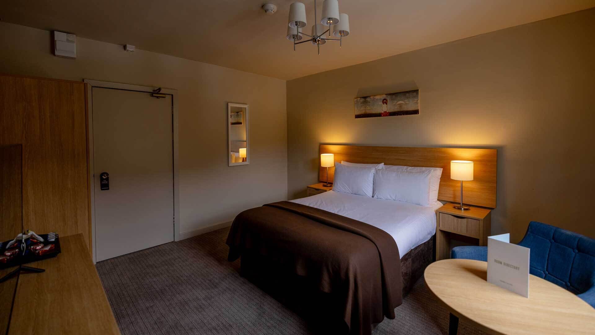 A hotel room with a bed, bedside lamps, a desk with a chair, a wooden door, and a small table with a sign.
