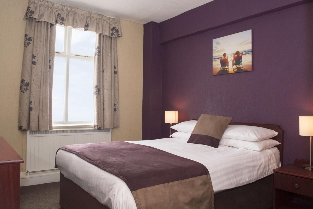 A hotel room with a bed, two bedside lamps, a window with curtains, a wall-mounted painting, and a radiator.