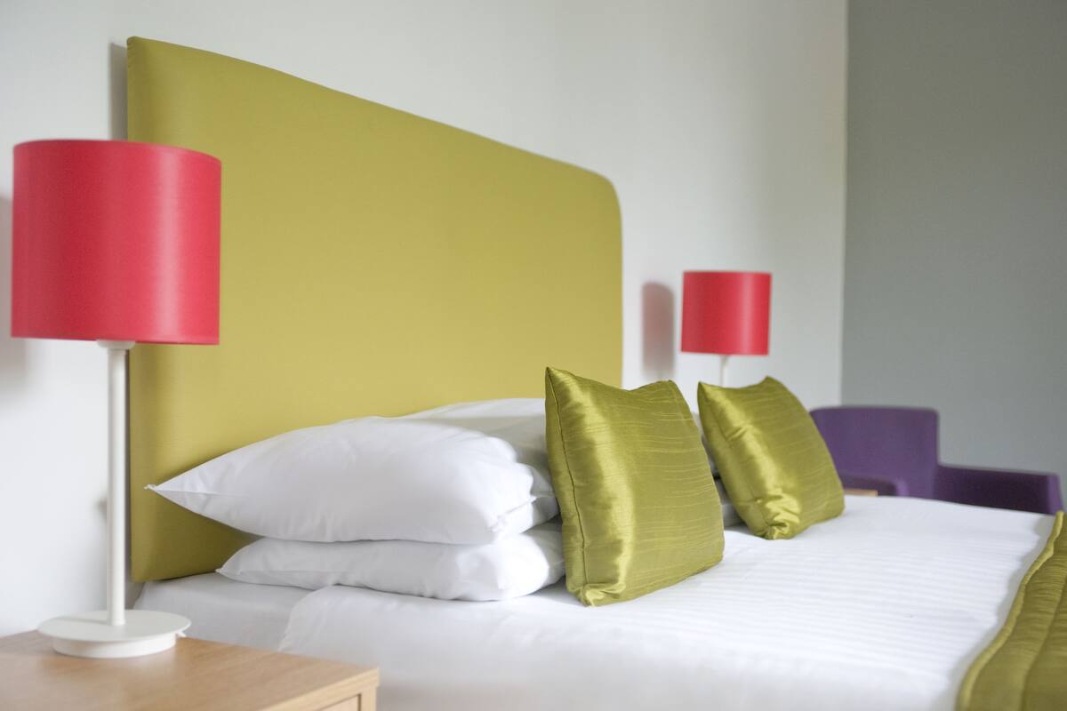 A neatly made bed with white and green pillows, a red lamp on a bedside table, and a purple chair in the background.