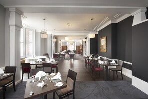 Un comedor amplio con varias mesas y sillas, ventanas grandes con persianas y una pared de acento oscura con un cuadro enmarcado.