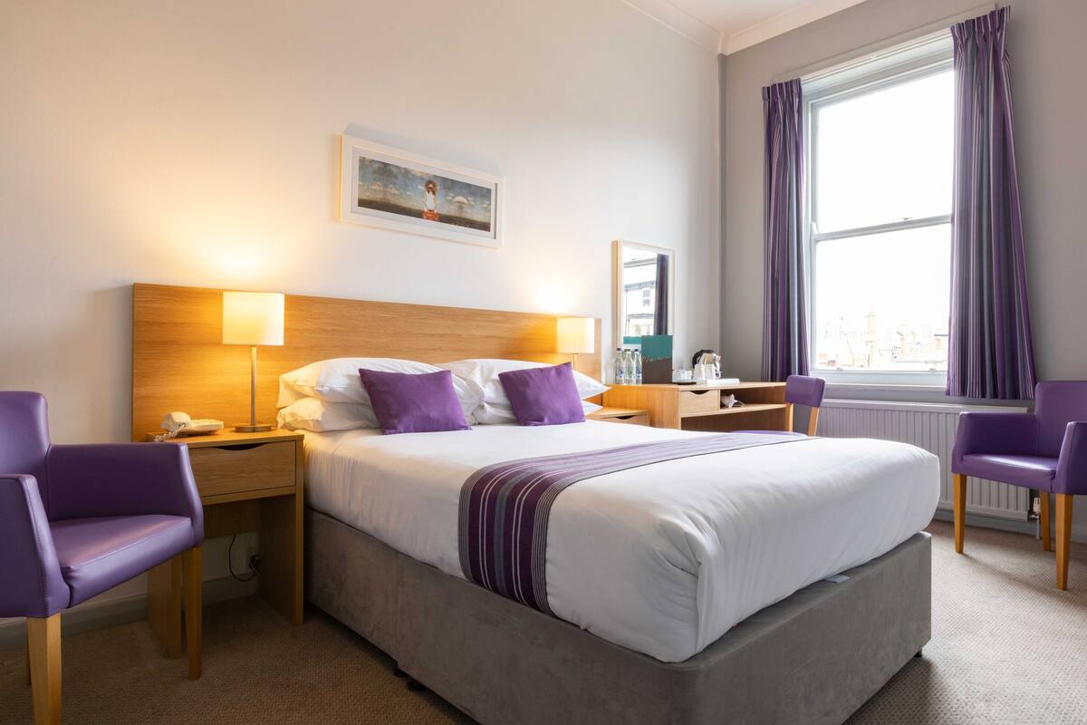 A hotel room with a large bed, two bedside tables with lamps, a chair, a desk, and a window with curtains.