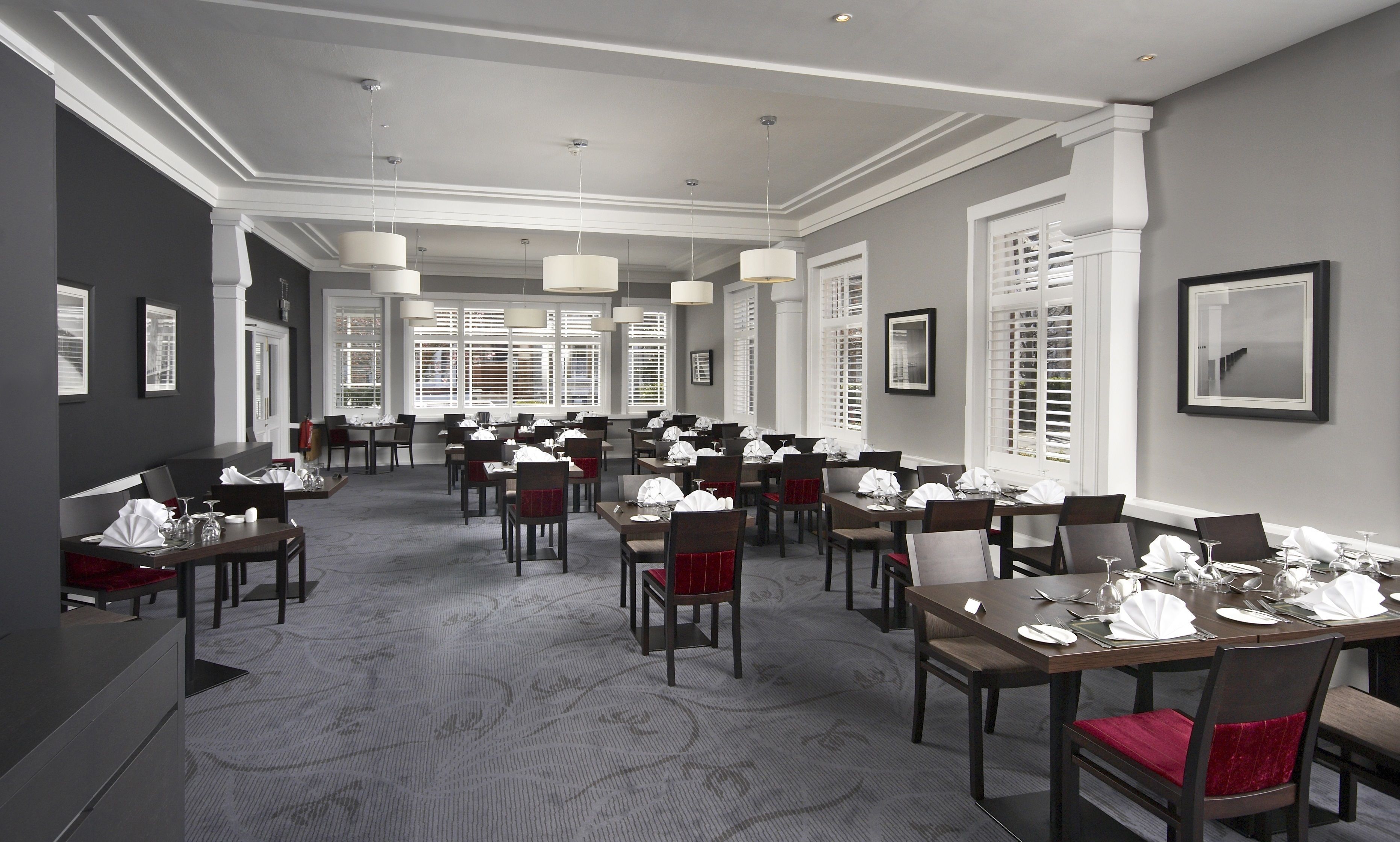 A hotel dining area with multiple tables set for service, large windows with blinds, and a carpeted floor.