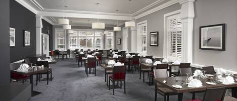 A hotel dining area with multiple tables set for service, large windows with blinds, and a carpeted floor.