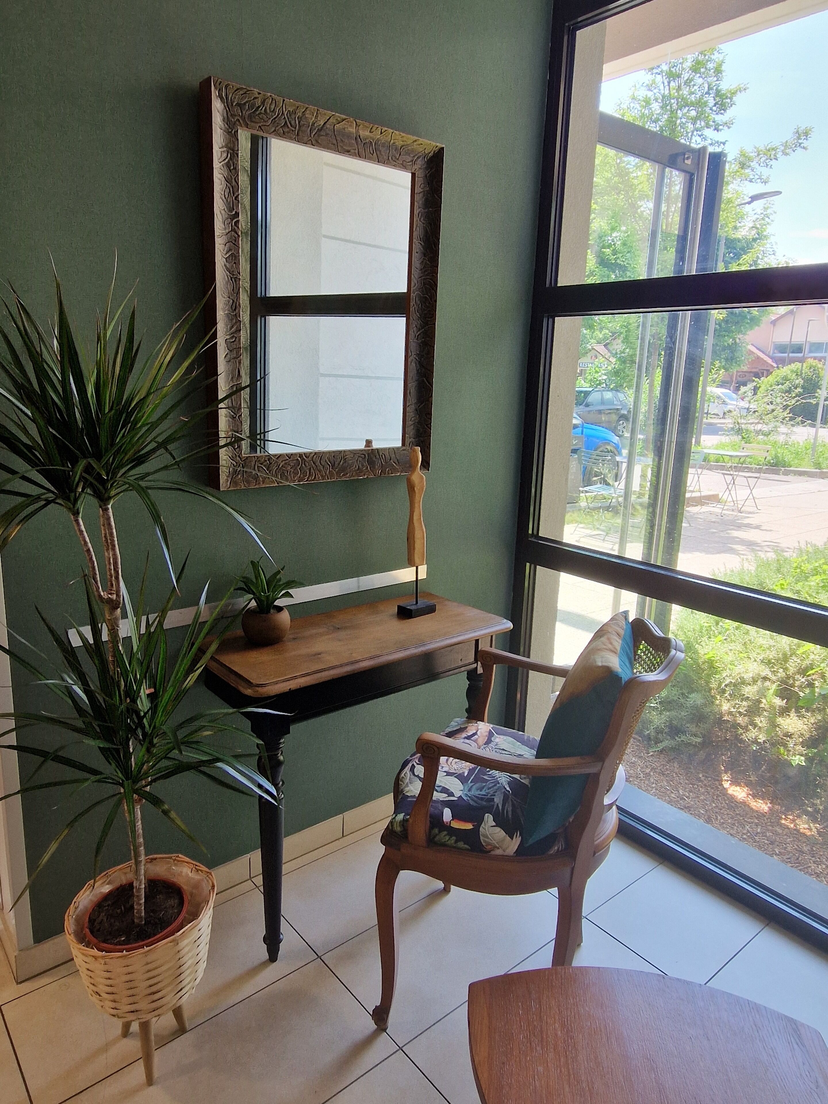 Ein Zimmer mit einem Holztisch, einem Stuhl mit Sitzkissen, einem Blumentopf und einem Spiegel an der Wand.