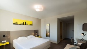 Een hotelkamer met een bed, een bureau met een laptop, een stoel, een nachtkastje met een lamp, een spiegel en een stadsgezicht aan de muur.