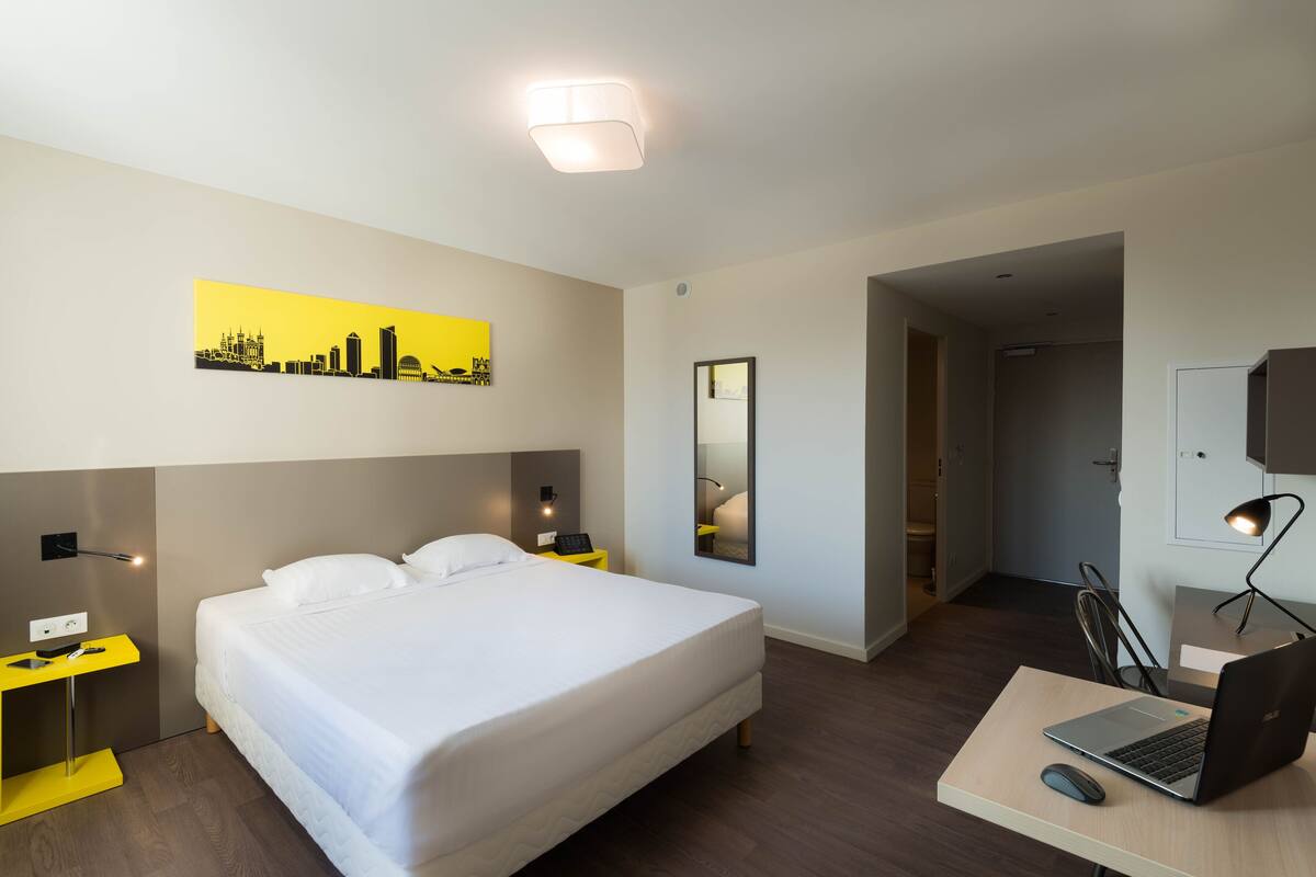 A hotel room with a bed, a desk with a laptop, a chair, a bedside table with a lamp, a mirror, and a cityscape artwork on the wall.