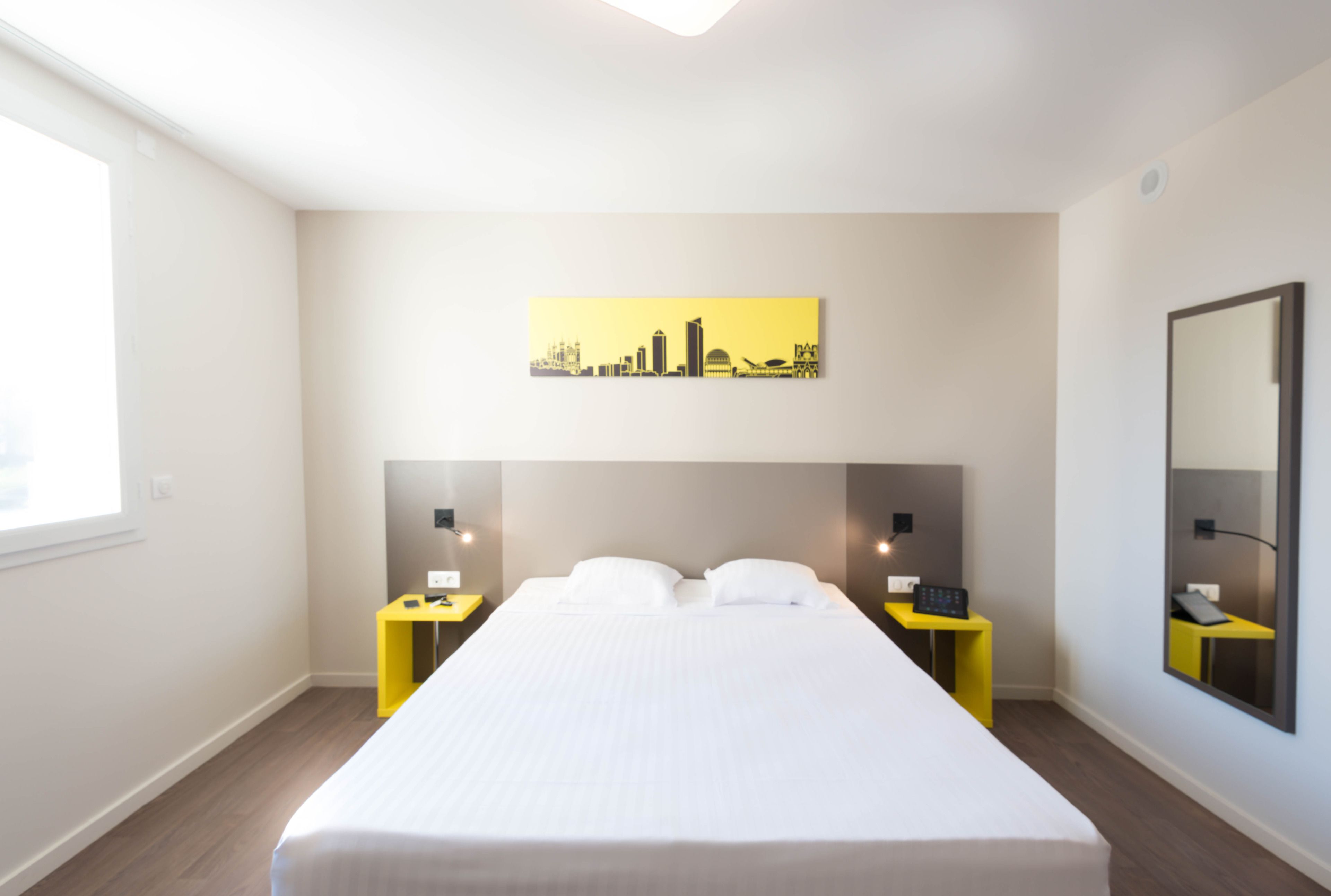 Habitació d'hotel amb un llit gran, dues taules de nit, un quadre de paisatge urbà, un mirall i una finestra.