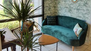 Een kamer met een smaragdgroene bank, een houten bijzettafeltje en een groot raam met uitzicht op groen.