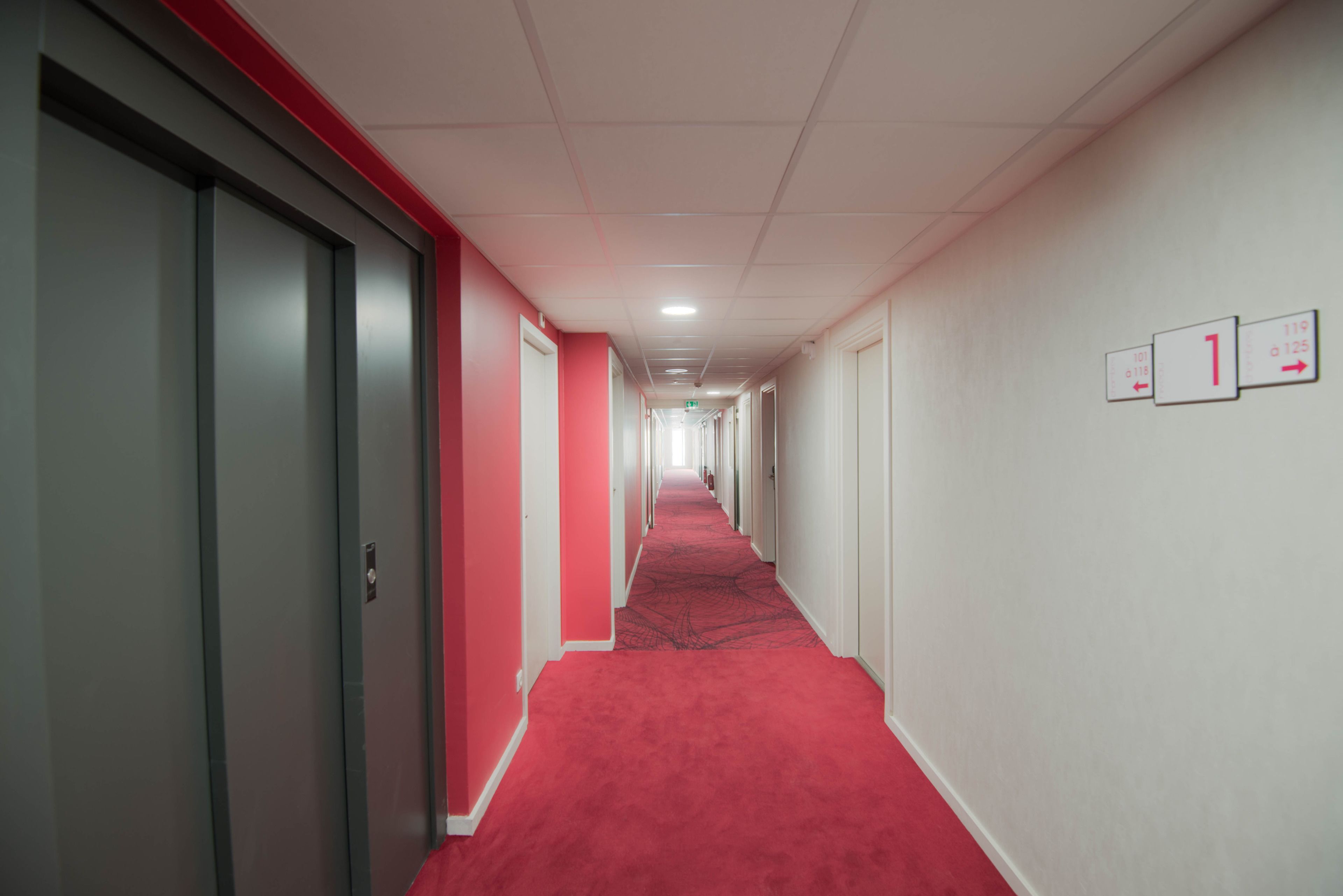 A long, narrow hallway with a red carpet, white walls, and doors on both sides.