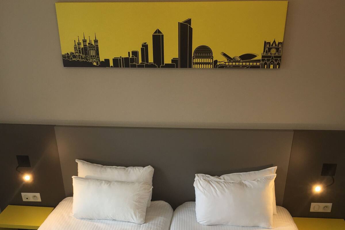 A hotel room with two single beds, each with white linens and pillows, and a city skyline artwork above the beds.