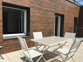 Un espacio exterior con mesas y sillas blancas, un muro de madera y una ventana.