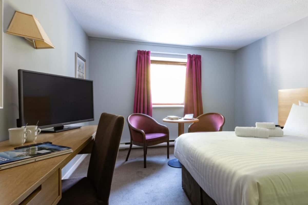 A hotel room with a bed, a desk with a TV, a chair, a small table, and a window with curtains.