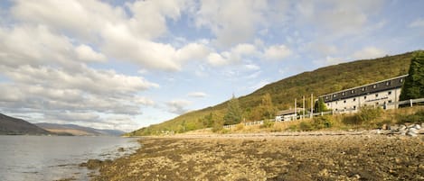 A coastal hotel with a rocky shoreline, a mountain backdrop, and a clear sky.