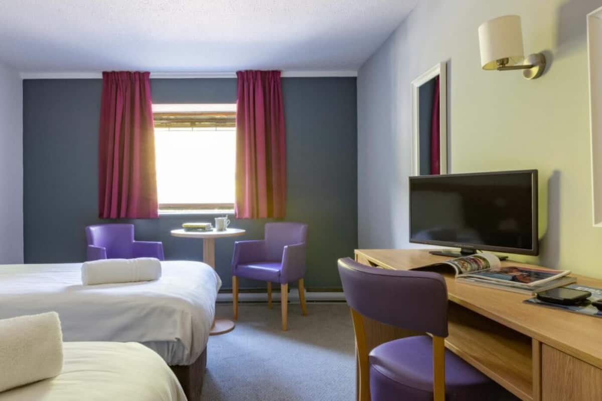 A hotel room with two beds, a desk with a TV, a chair, and a window with curtains.