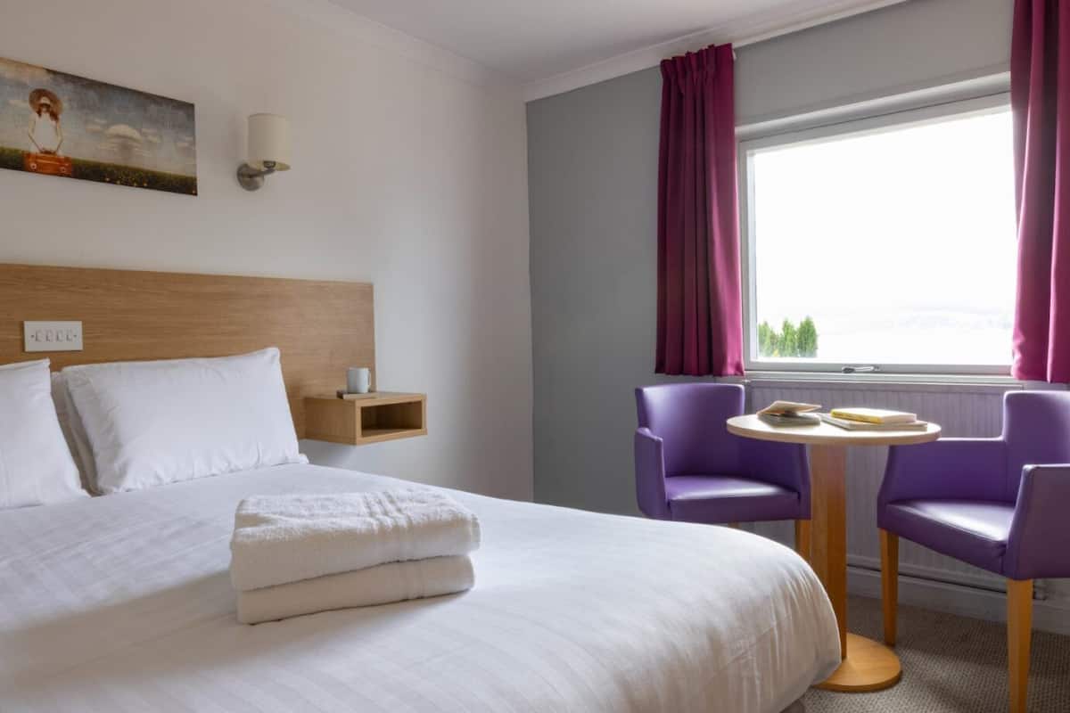 A hotel room with a bed, a nightstand, a table with chairs, and a window with curtains.