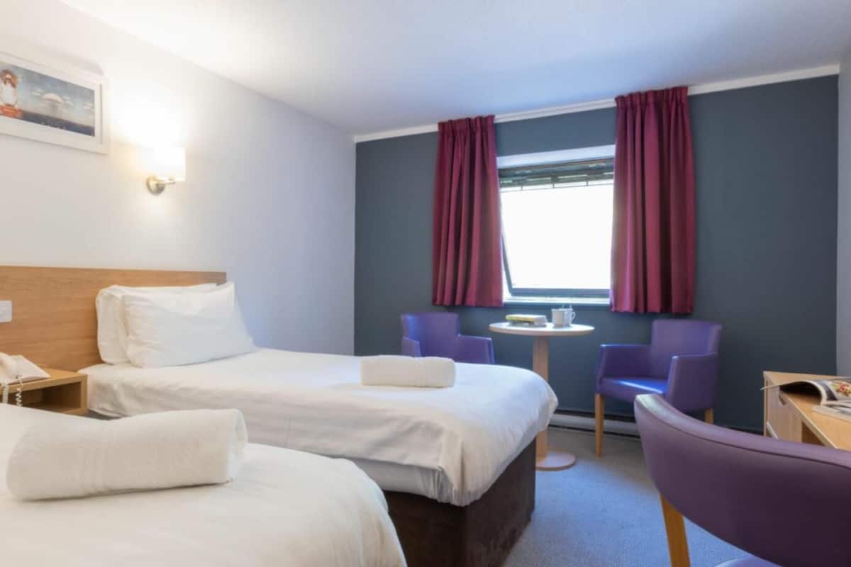 A hotel room with two beds, a desk, a chair, a small table, and a window with curtains.