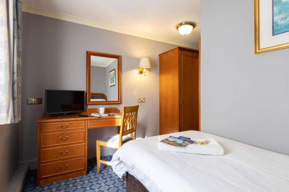 A hotel room with a bed, a desk with a computer, a chair, a wardrobe, and a mirror.