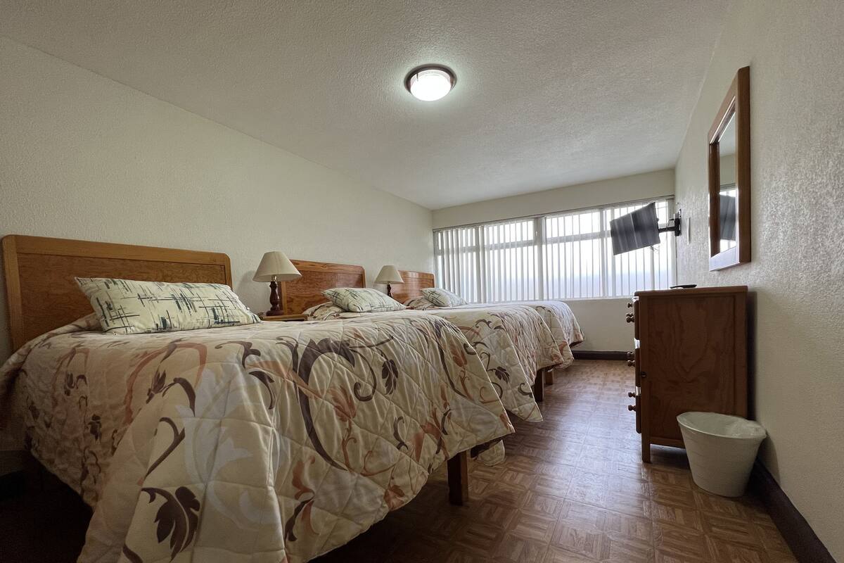 A bedroom with two beds, wooden headboards, a wooden dresser, a TV, and a window with blinds.