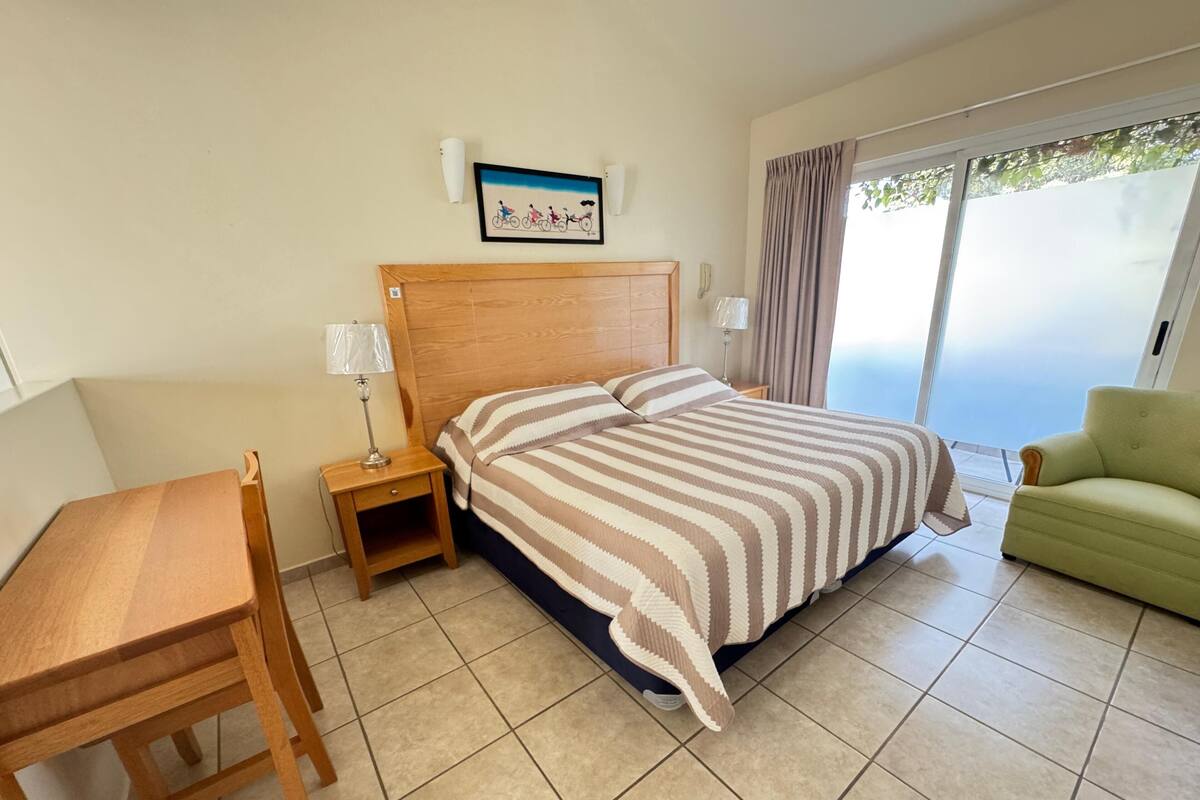 A hotel room with a bed, a wooden nightstand, a lamp, a green armchair, and a window with curtains.