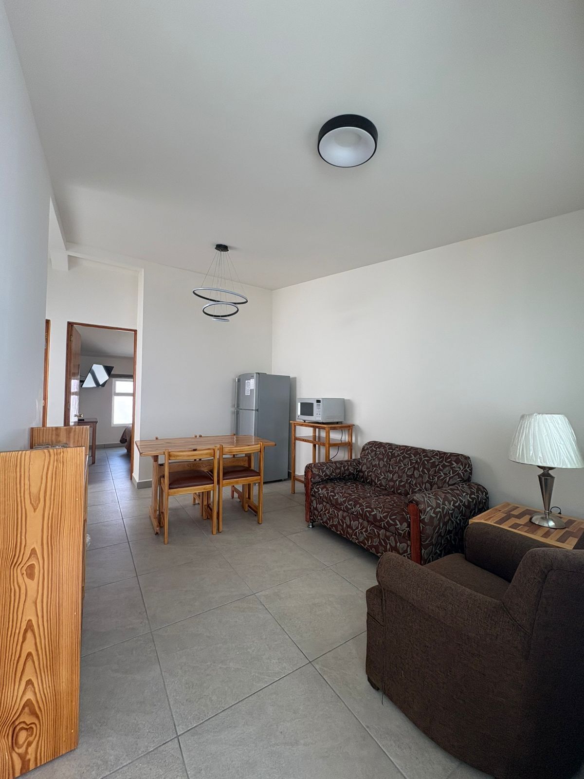 A living room with a sofa, a dining table, and a kitchen area with a refrigerator and microwave.