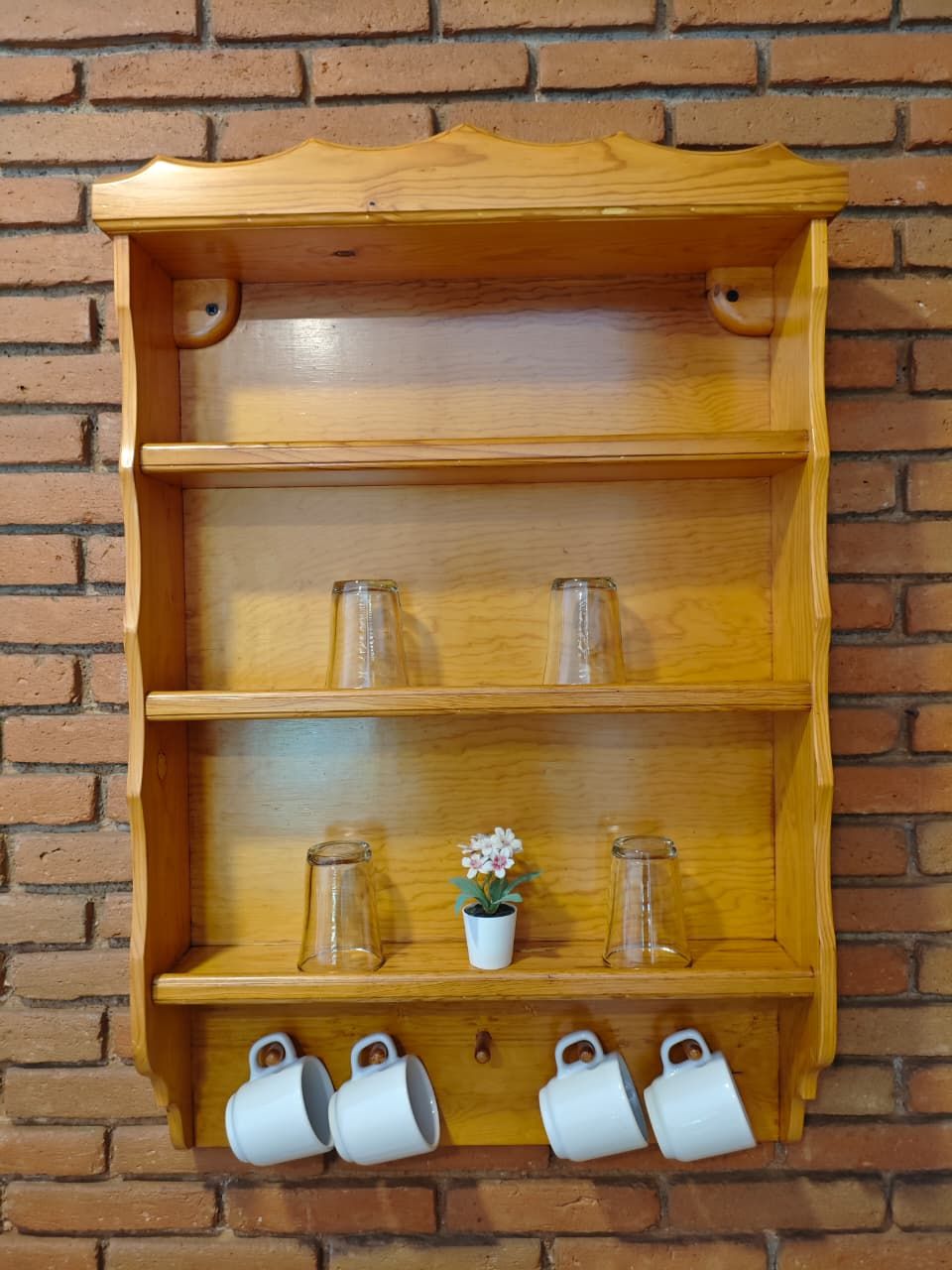 A wooden shelf with three glass bottles, a small potted plant, and four white mugs against a brick wall.