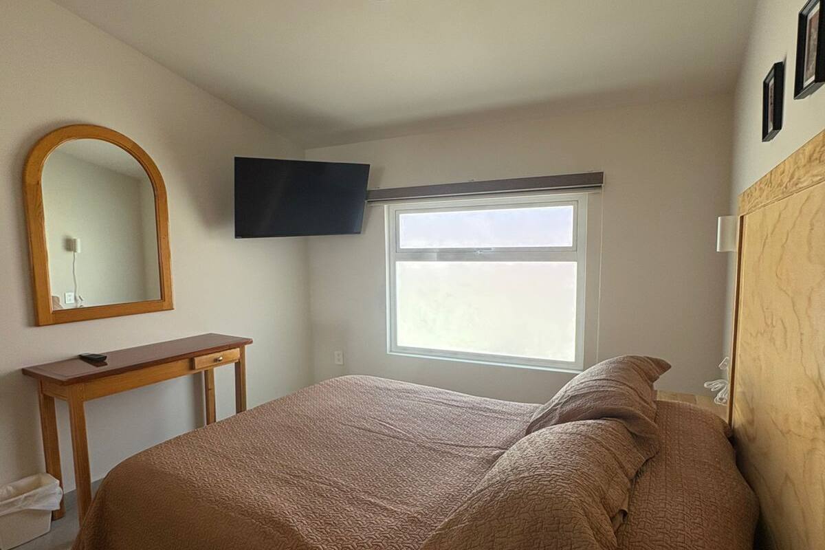 A bedroom with a bed, a wooden bedside table, a mirror, a flat-screen TV mounted on the wall, and a window with a view.