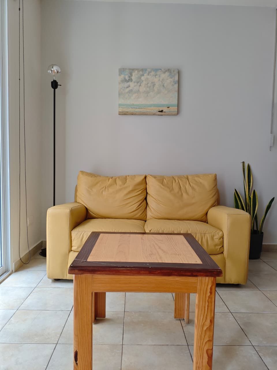 A room with a yellow sofa, a wooden table, a painting of a beach scene, a floor lamp, and a potted plant.