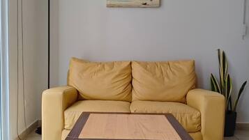 A room with a yellow sofa, a wooden table, a painting of a beach scene, a floor lamp, and a potted plant.