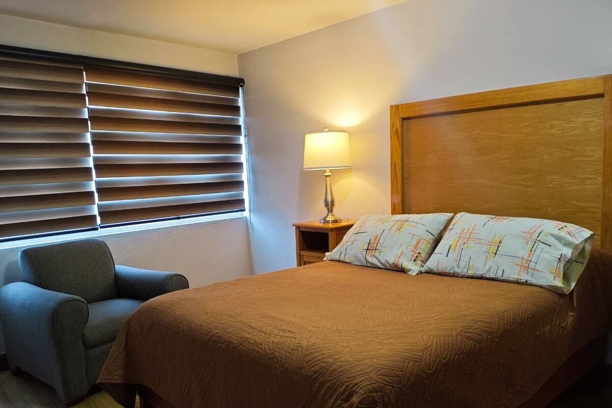 A bedroom with a large bed, a bedside table with a lamp, a chair, and a window with blinds.