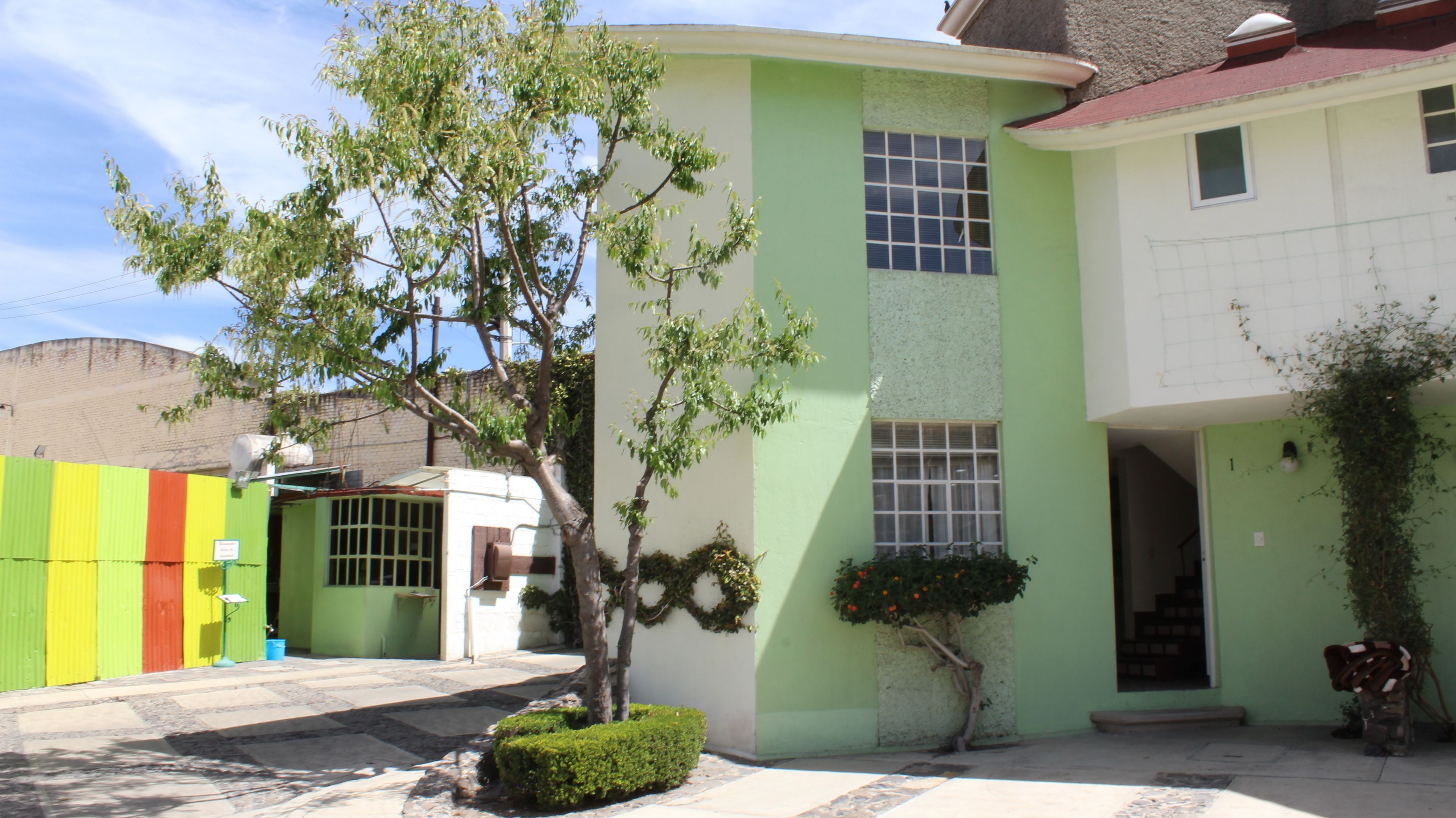 A two-story building with a green facade, a small tree in front, and a colorful fence in the background.