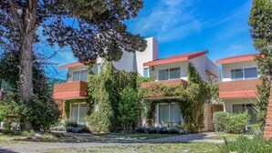 A modern multi-story building with a brick facade, large windows, and a well-maintained garden with various plants and a paved driveway. - Privada 400 Casas & Suites (Pachuca)