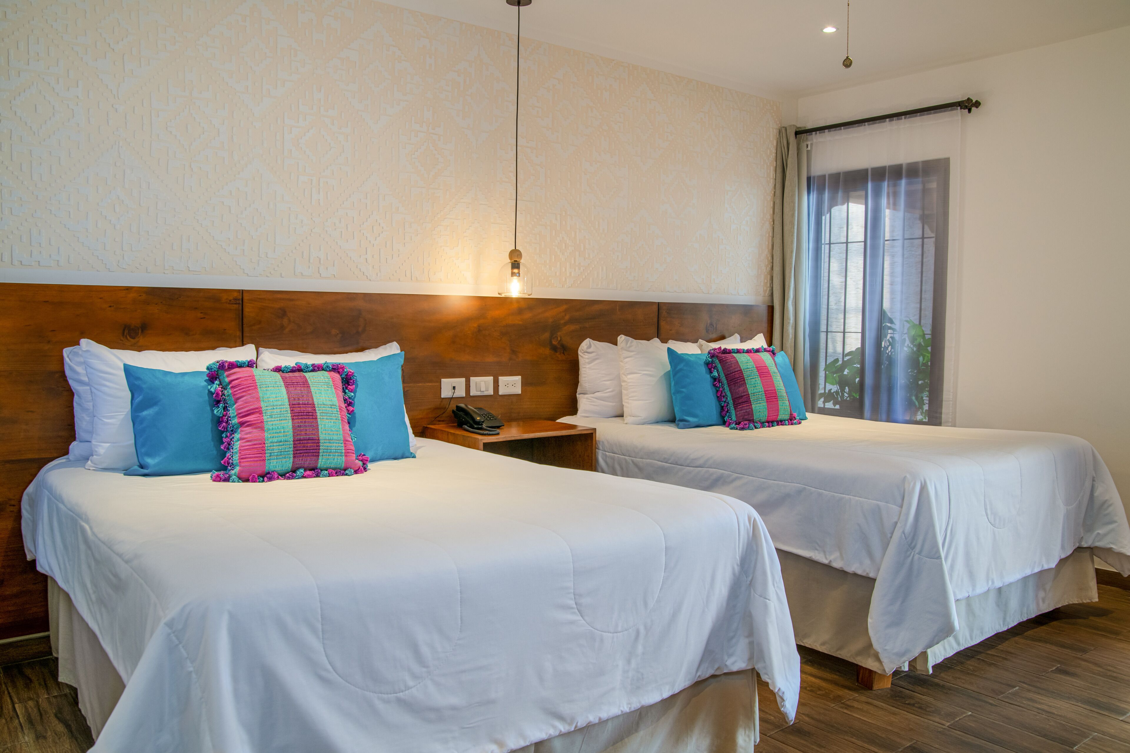 Une chambre d’hôtel avec deux lits, chacun agrémenté de draps blancs et d’oreillers colorés. On y trouve une tête de lit en bois, une table de chevet avec un téléphone et une fenêtre avec des rideaux.