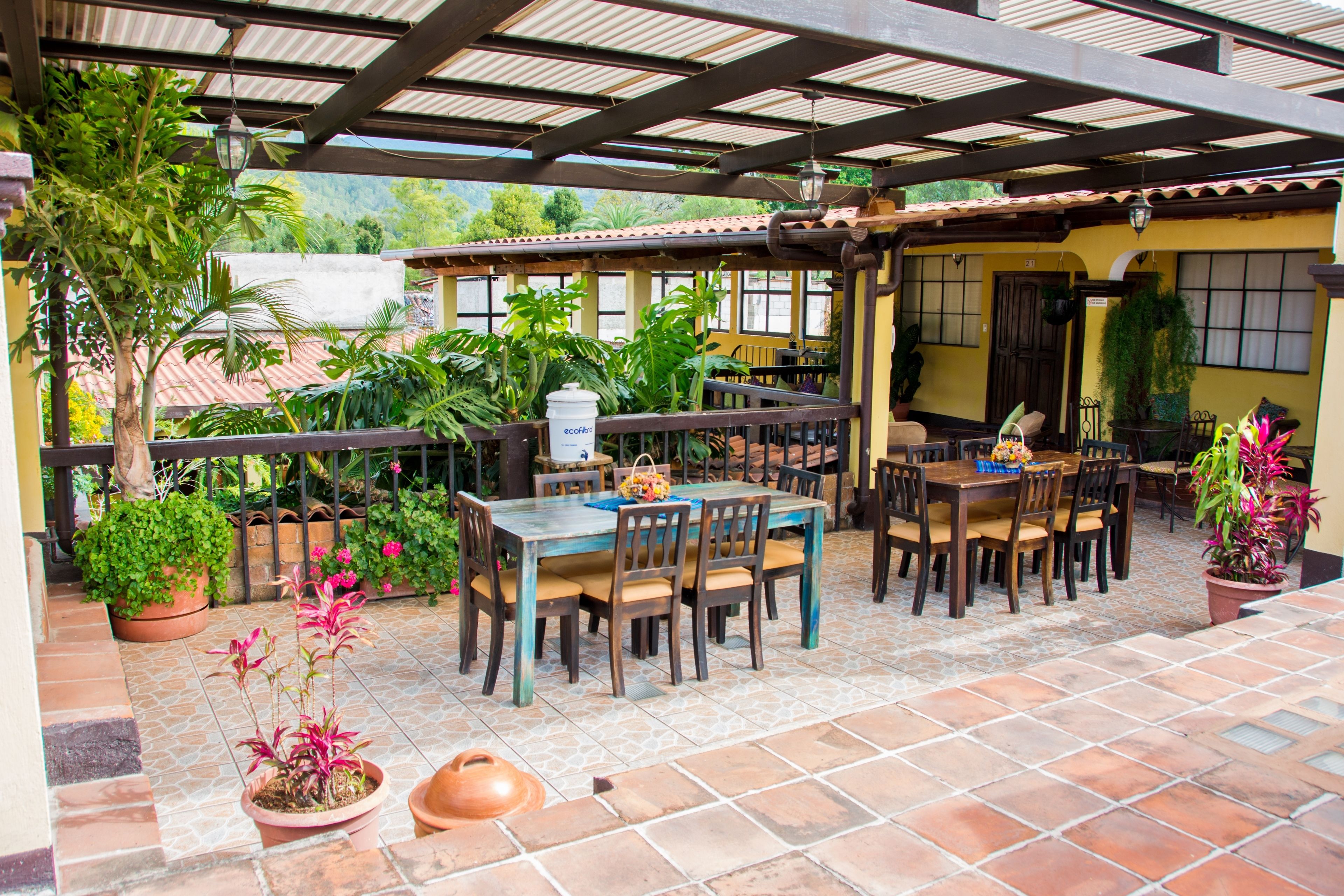 Un patio al aire libre con una mesa y sillas cubiertas para comer, rodeado de plantas en macetas y un suelo de baldosas.