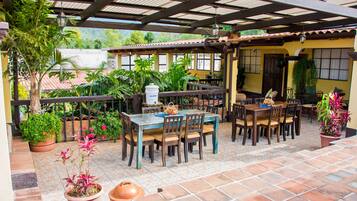 Un patio al aire libre con una mesa y sillas cubiertas para comer, rodeado de plantas en macetas y un suelo de baldosas.