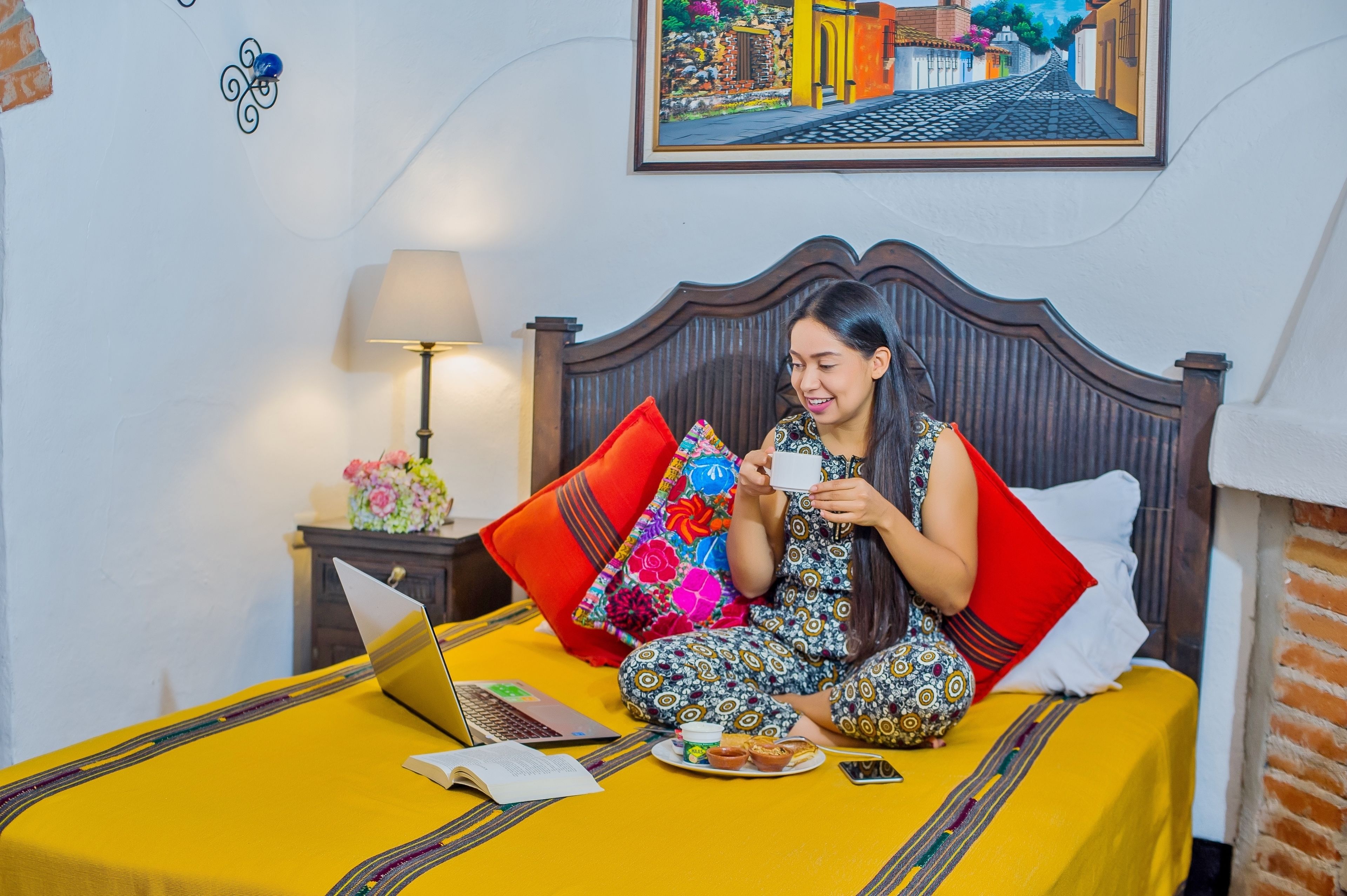 Una cama con una colcha amarilla, una computadora portátil, un libro y un plato de comida.