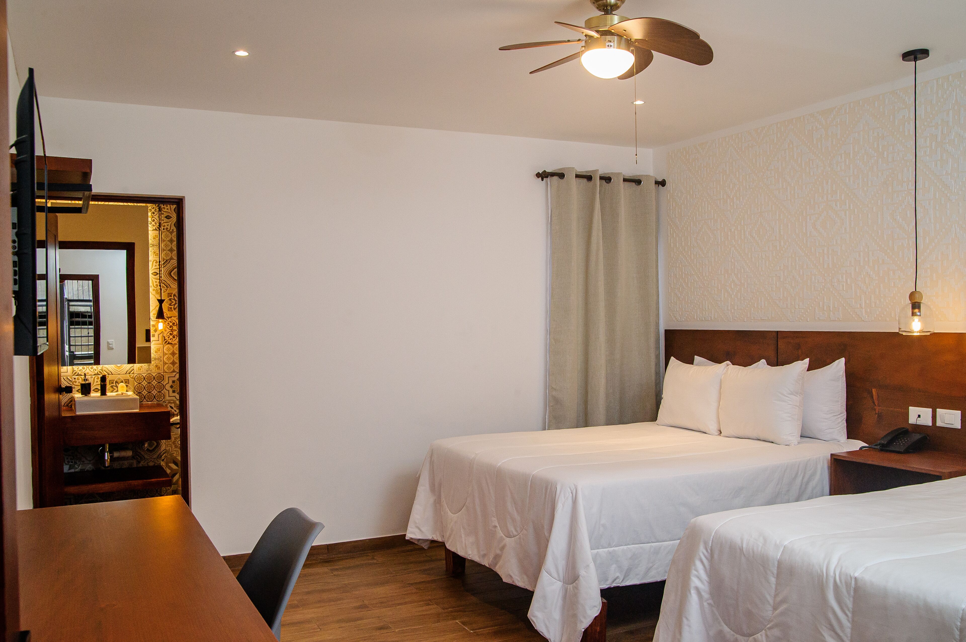 A hotel room with two single beds, a wooden desk with a chair, a small bathroom with a mirror, and a ceiling fan.