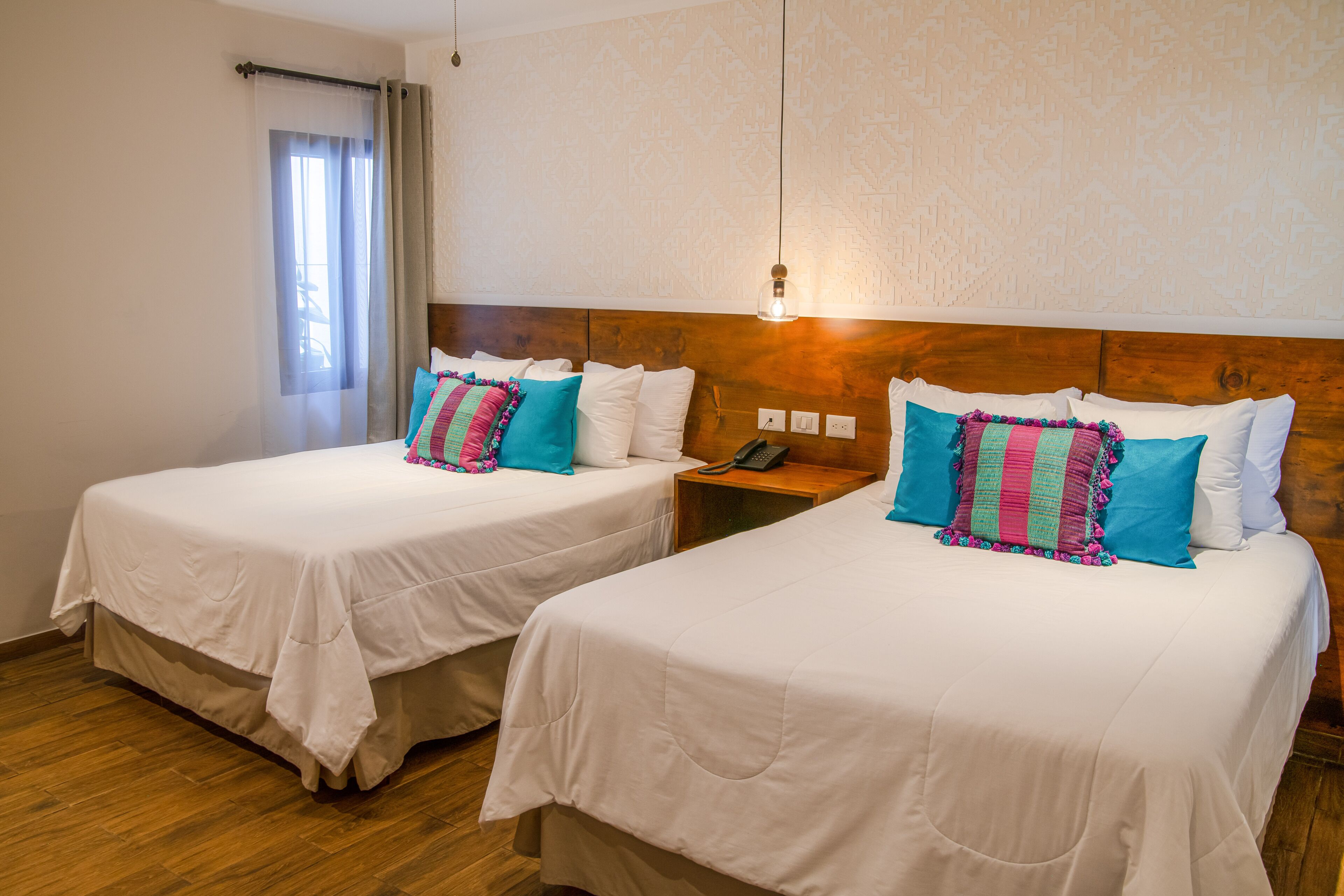 Dos camas individuales con ropa blanca y almohadas de colores, un cabecero de madera, una mesita de noche con un teléfono y una ventana con cortinas traslúcidas.