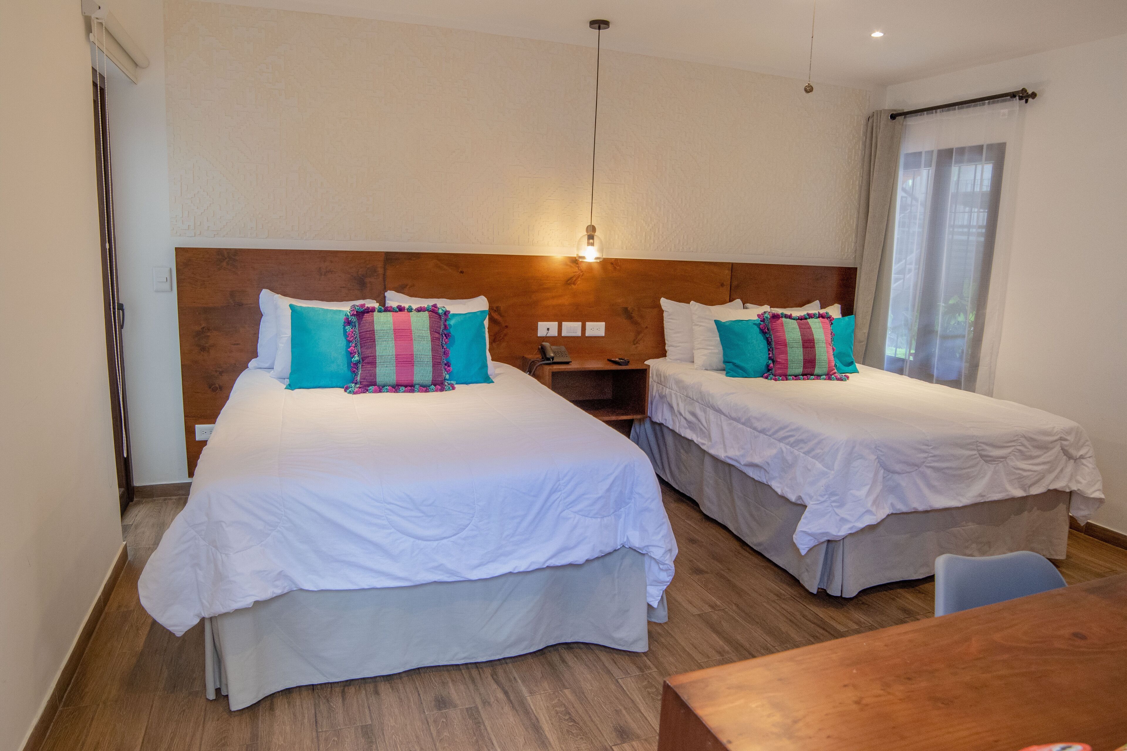 A hotel room with two beds, wooden headboards, a wooden table, and a small desk with a telephone.