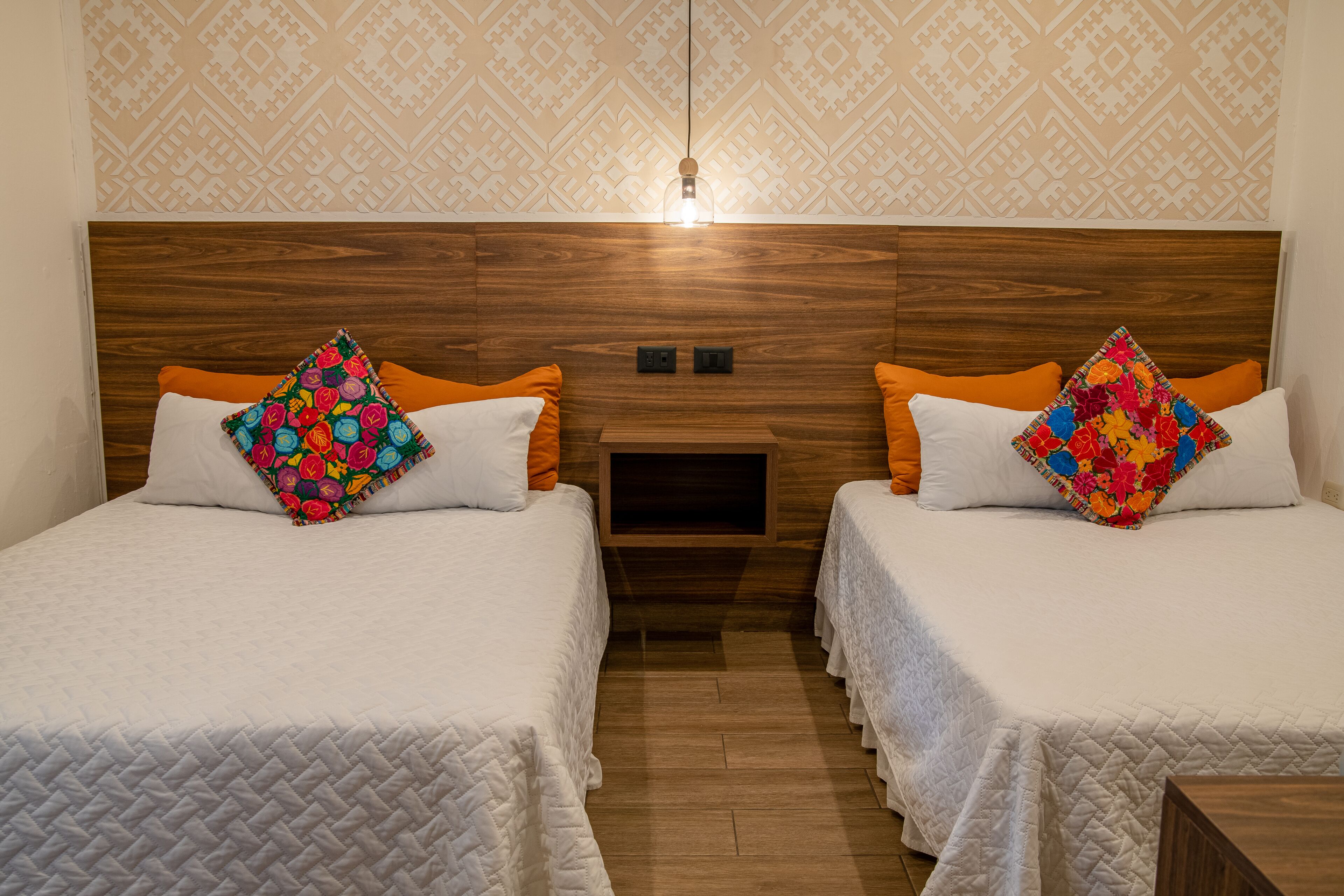 A hotel room with two beds, each with a white quilted bedspread and colorful floral pillows. There is a wooden headboard, a small bedside table, and a wall-mounted light fixture.