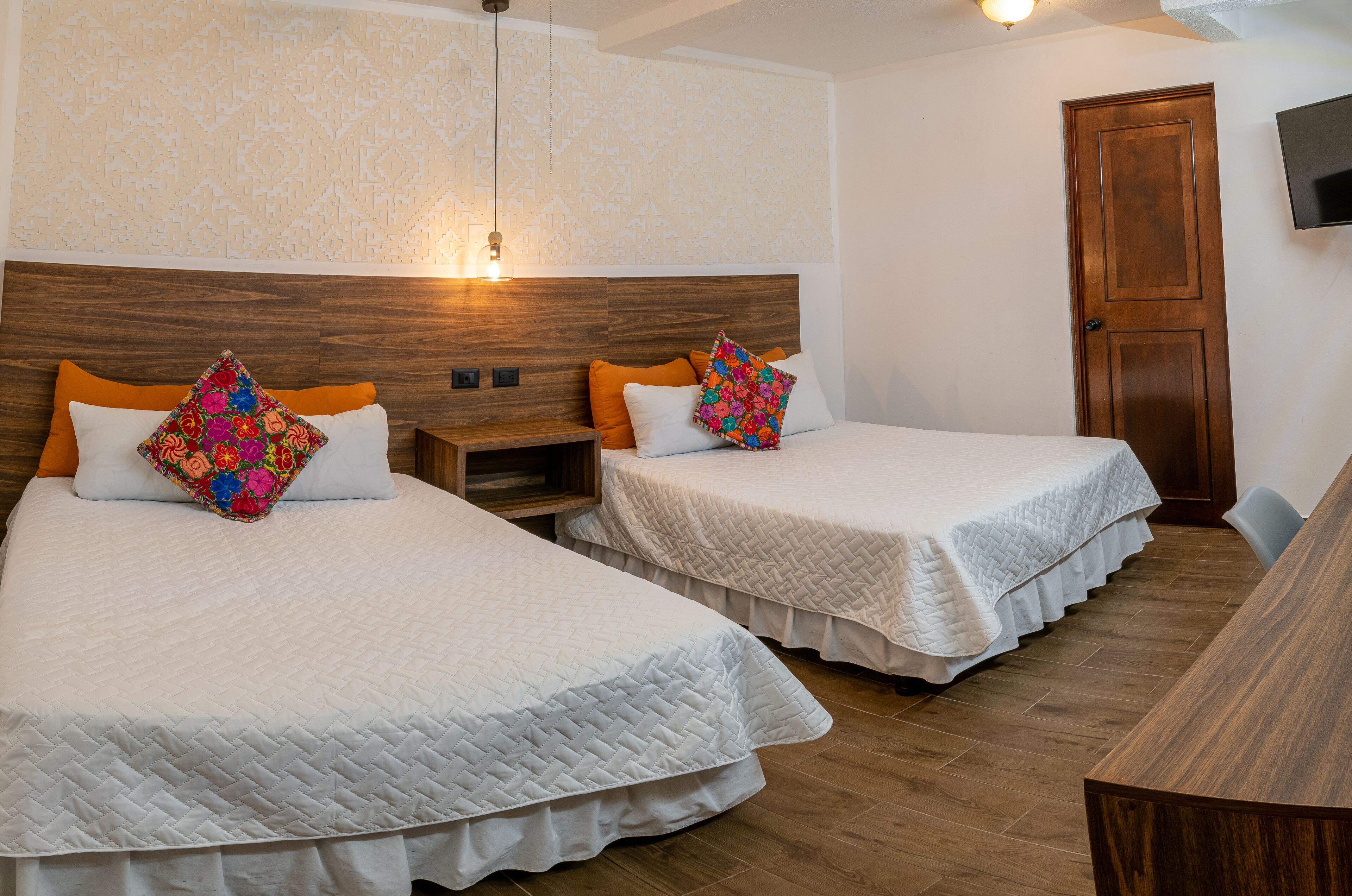 Habitación de hotel con dos camas, cada una con colcha blanca acolchada y almohadas naranjas. Se observa un cabecero de madera, una mesita de noche y un televisor montado en la pared.