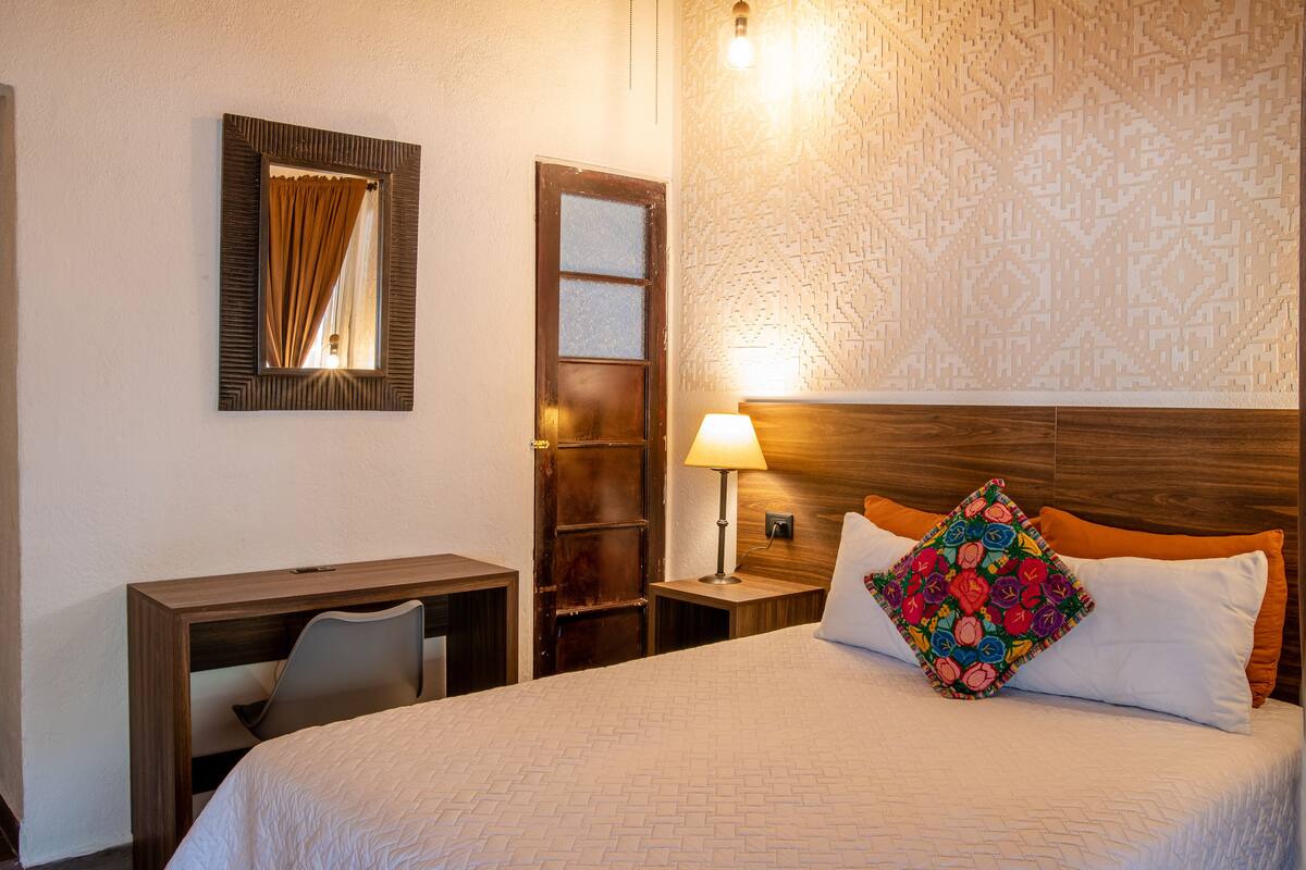 A hotel room with a bed, a desk, a chair, a nightstand with a lamp, a mirror, and a door.