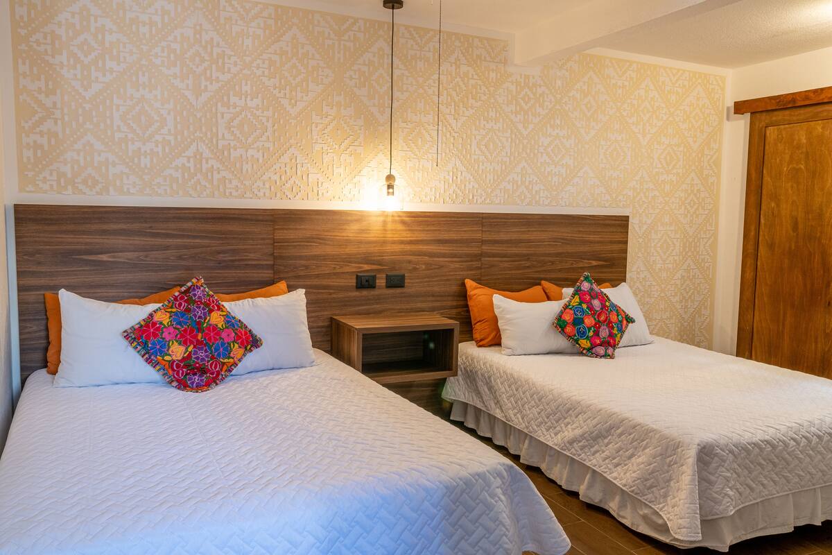 A hotel room with two beds, each with a wooden headboard, white bedding, and colorful pillows. There is a nightstand between the beds and a wooden wardrobe to the right.