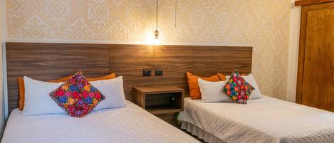 Habitación de hotel con dos camas, cada una con cabecera de madera, ropa de cama blanca y almohadas de colores. Hay una mesita de noche entre las camas y un armario de madera a la derecha.