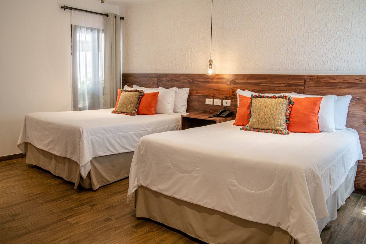 Two single beds with white bedding and orange pillows, wooden headboards, a wooden nightstand with a phone, and a wall-mounted light fixture.