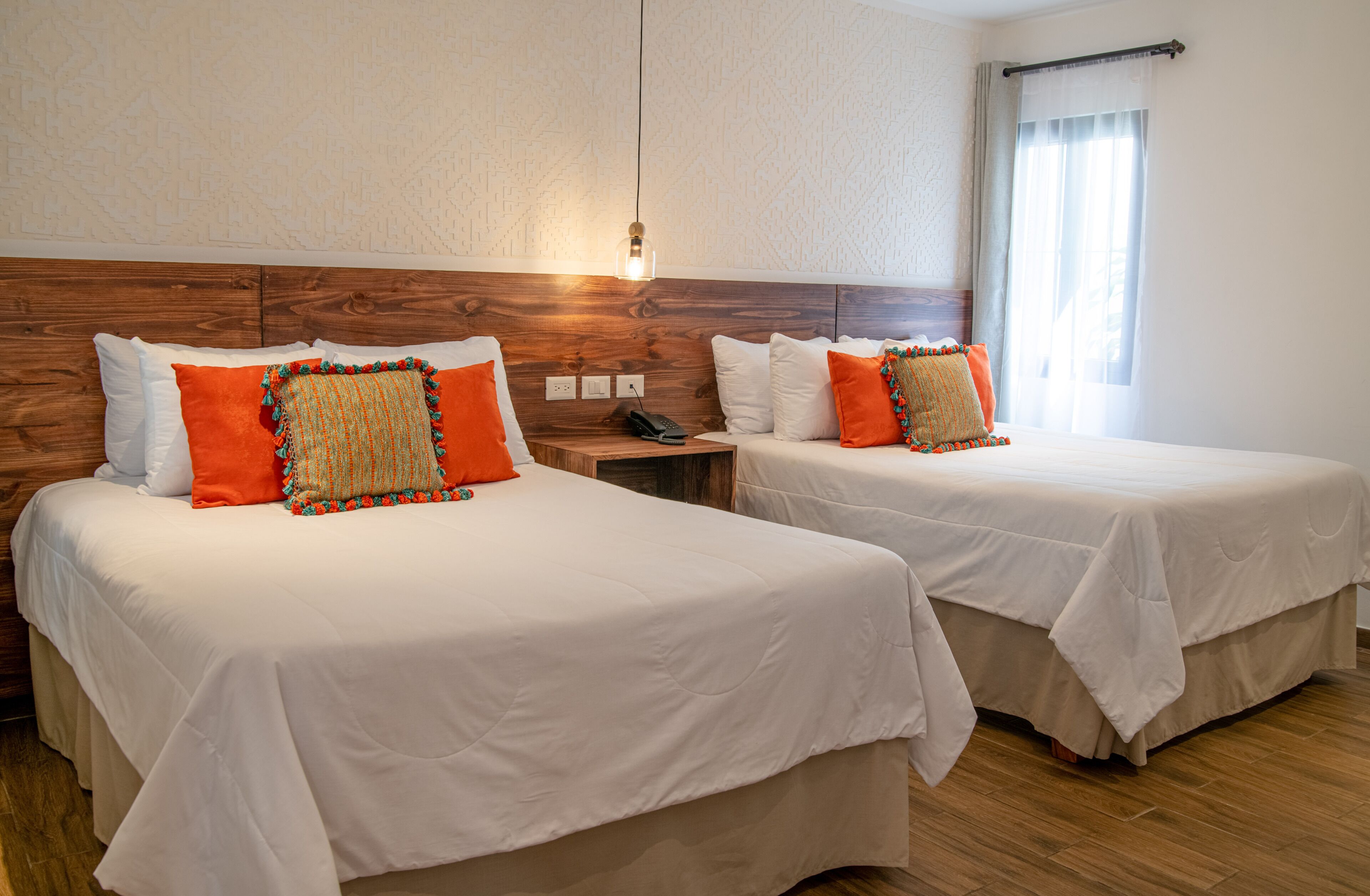 Two beds with white bedding and orange pillows, a wooden headboard, a bedside table with a phone, and a window with sheer curtains.