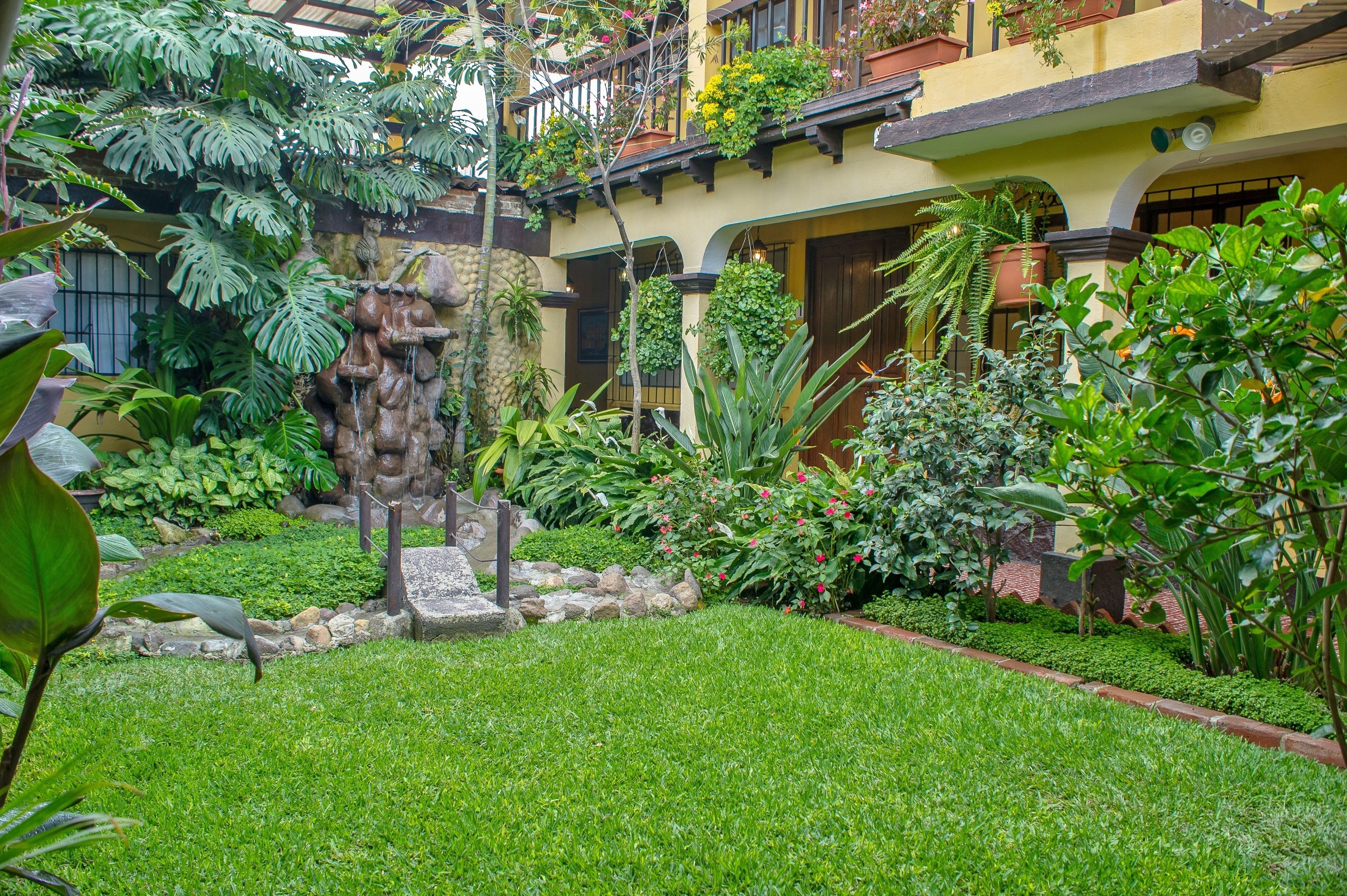 A lush garden with a stone fountain, various plants, and a yellow building with balconies.