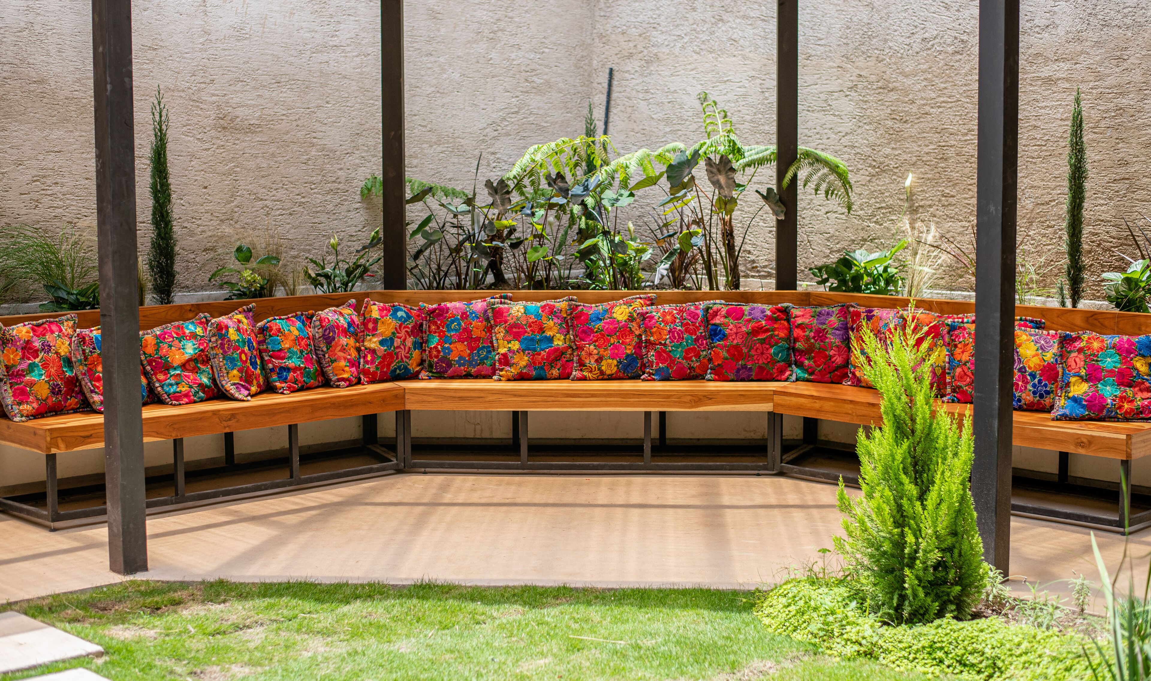 Un banco de madera curvo con cojines florales de colores, rodeado de plantas y una pared con textura.