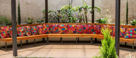 Un banco de madera curvo con cojines florales de colores, rodeado de plantas y una pared con textura.