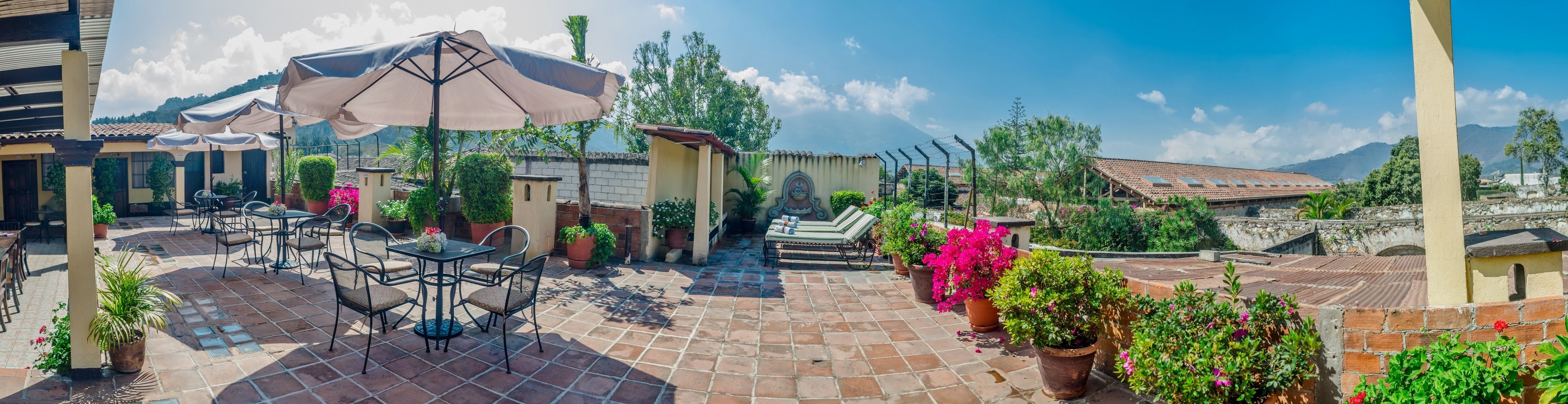 Un patio exterior con mesas y sillas, rodeado de plantas en macetas y vistas a la montaña.