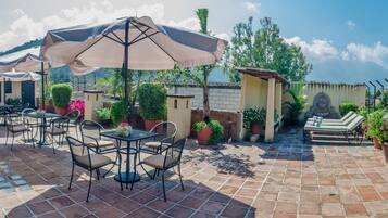 Un patio exterior con mesas y sillas, rodeado de plantas en macetas y vistas a la montaña.